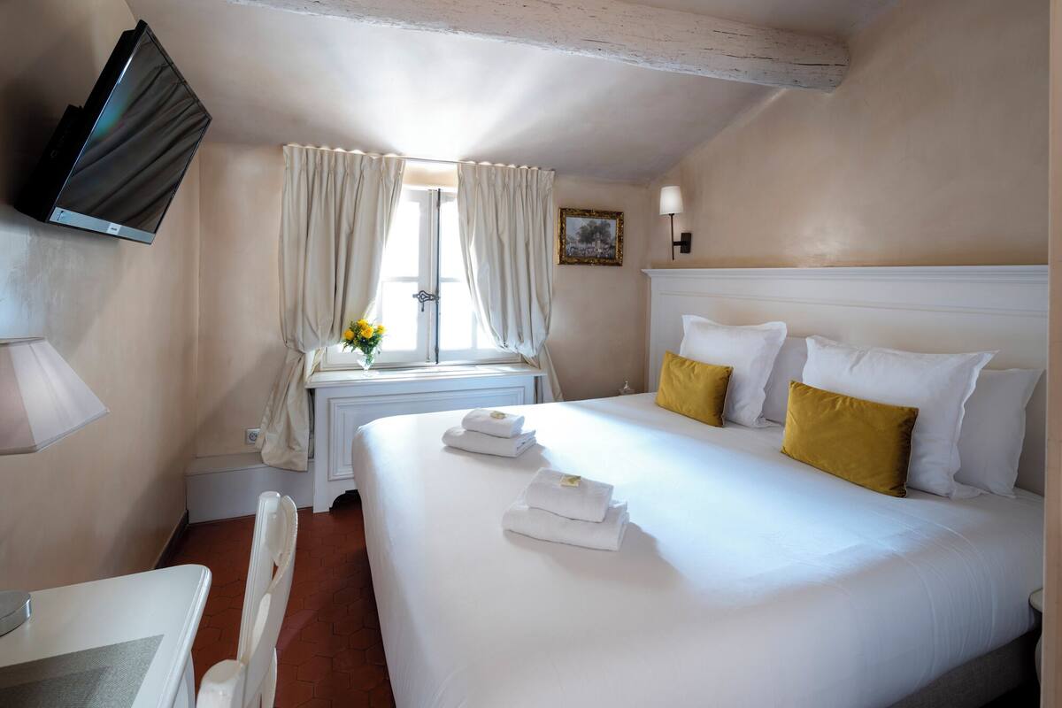 Una habitación de hotel con cama, escritorio, silla, televisor, ventana con cortinas y un cuadro en la pared.