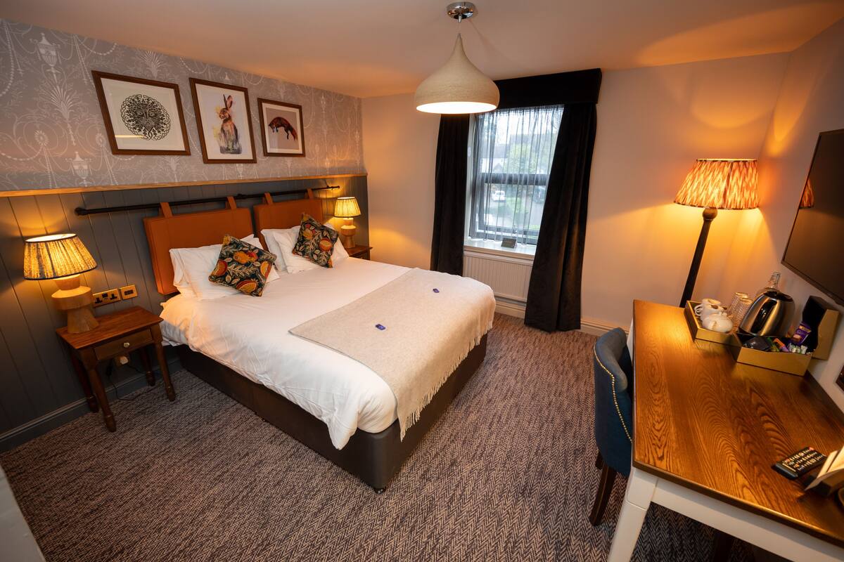 A hotel room with a bed, bedside lamps, a desk with a lamp, a window with curtains, and framed artwork on the wall.