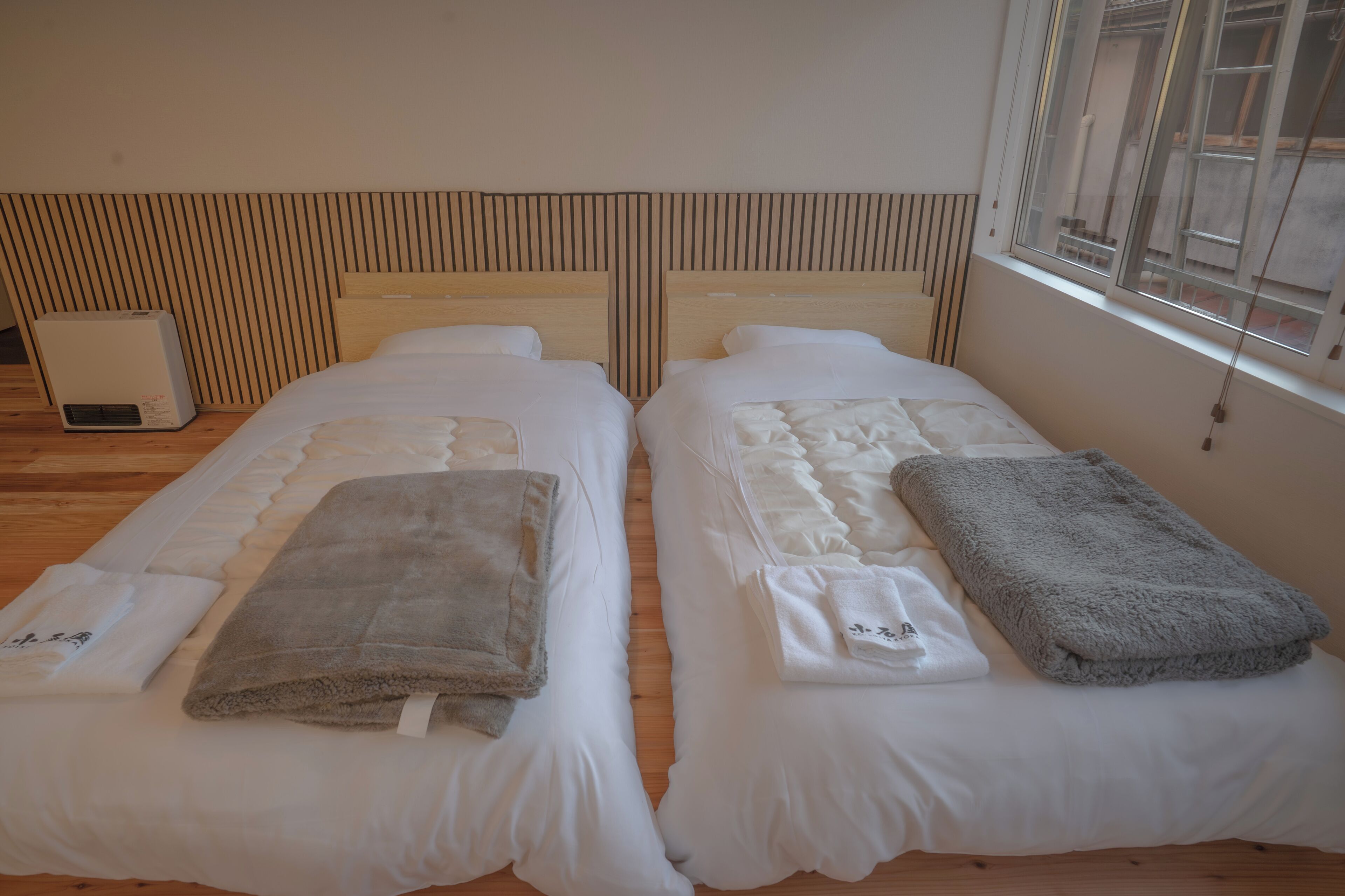 A room with two beds, each with a pillow and a blanket. There is a heater on the floor and a window with a view.