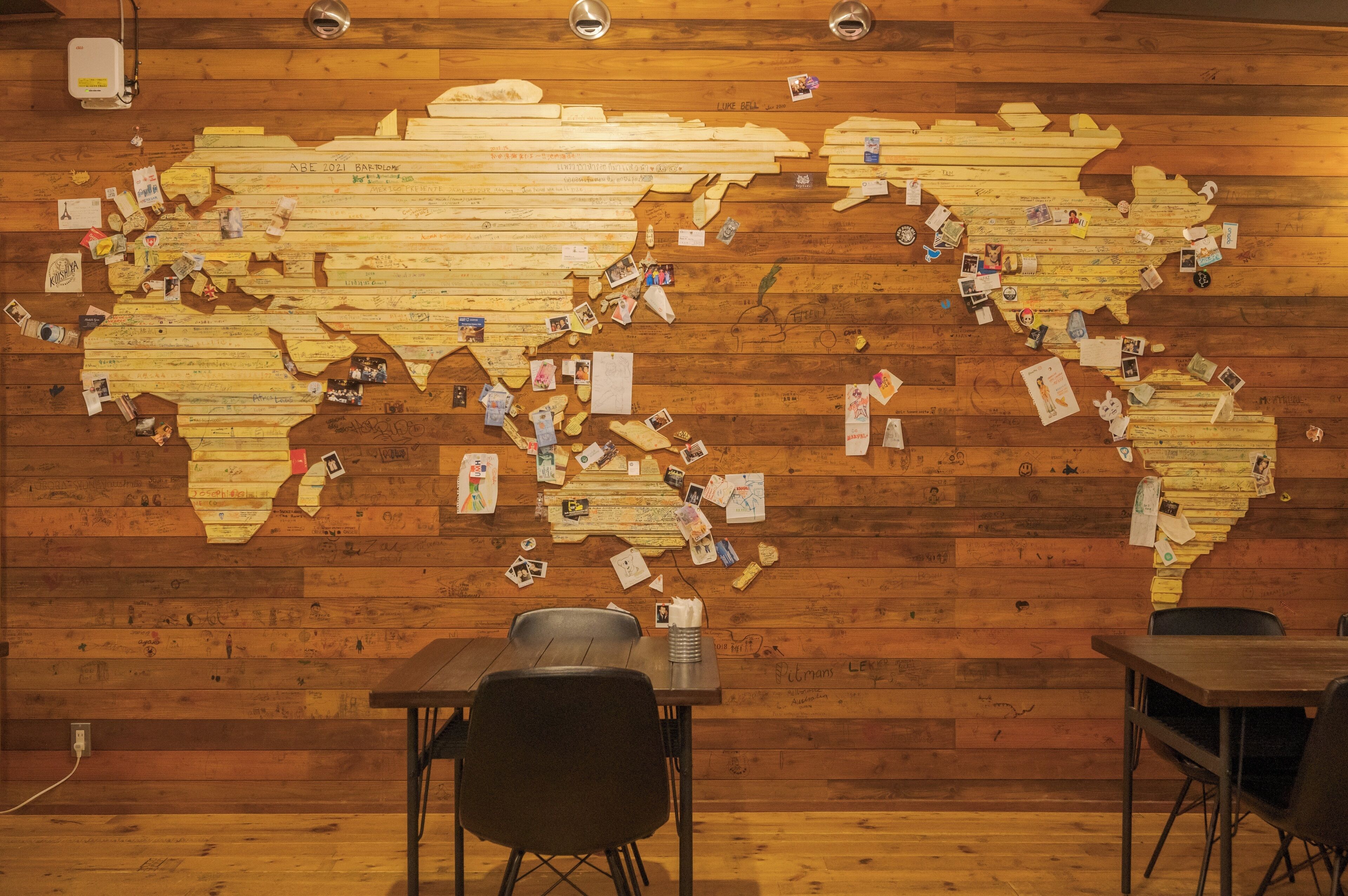 A wooden wall with a world map made of cut-out pieces, some with photos and notes. There are two tables and chairs in the foreground.