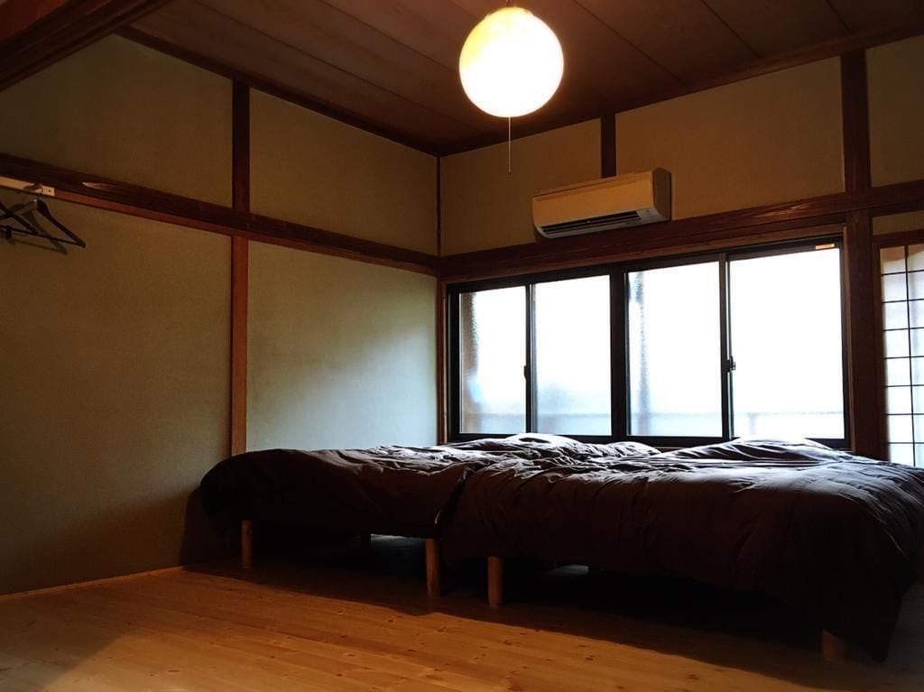 Una habitación con una cama, suelo de madera, una lámpara colgante y una ventana con persianas.