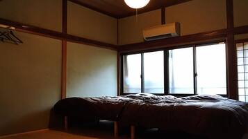 A room with a bed, wooden flooring, a hanging light fixture, and a window with blinds.