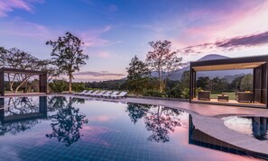 A pool with lounge chairs and a covered seating area, surrounded by trees and a mountainous backdrop.