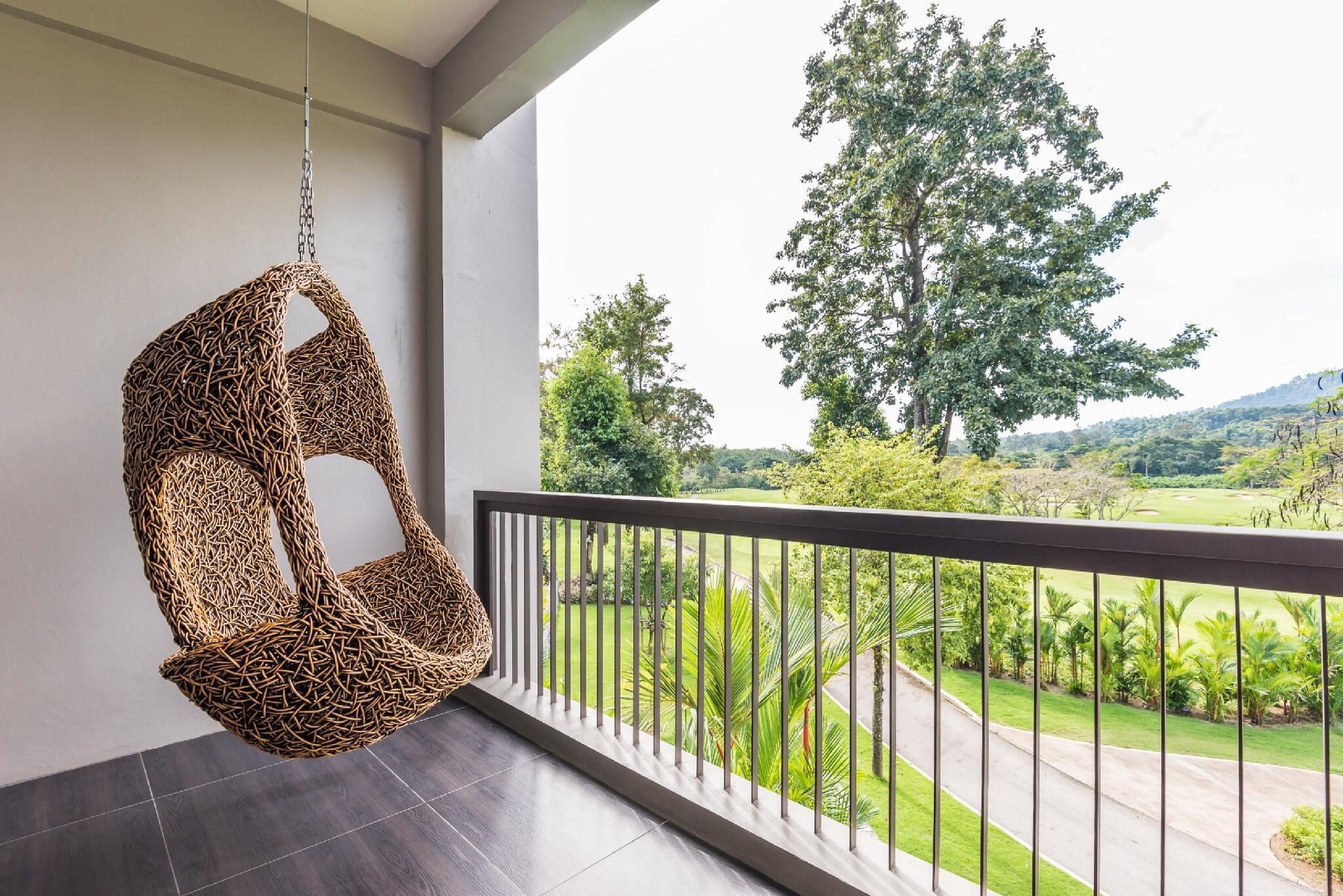 A wicker hanging chair on a balcony with a view of a lush green landscape and a road.