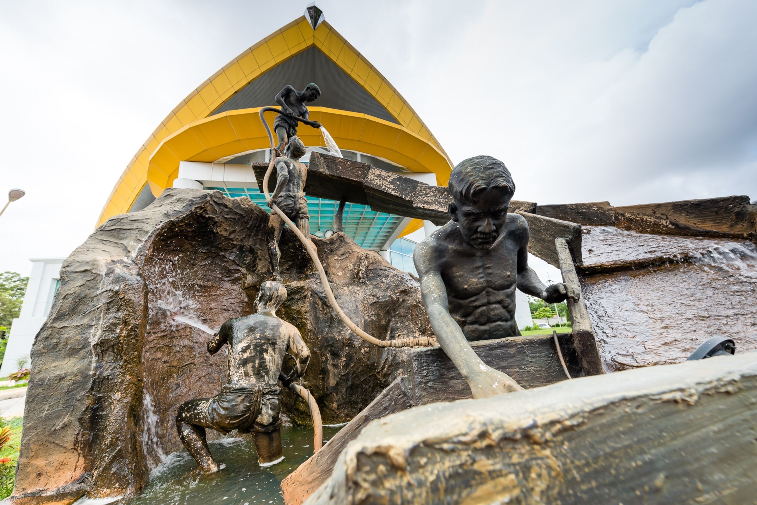A sculpture of children playing with water, a large rock, and a yellow building in the background.