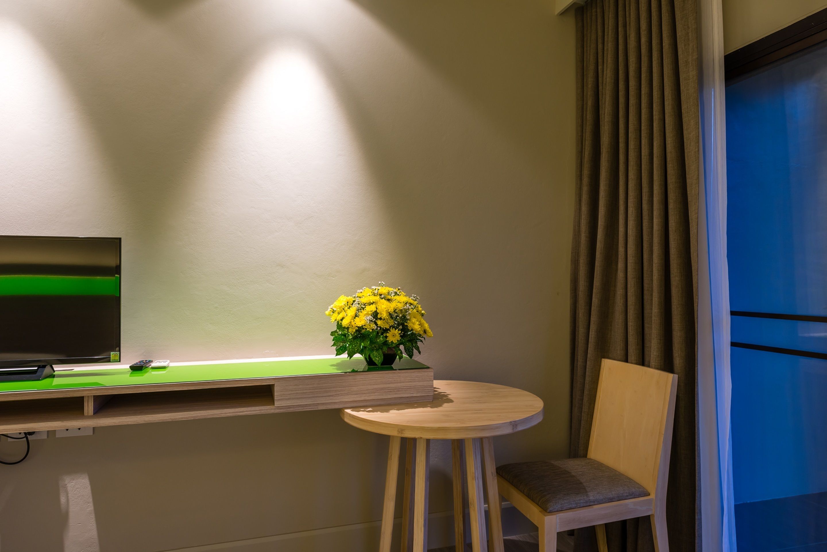 A hotel room with a TV on a green stand, a small table with a vase of yellow flowers, a wooden chair, and a window with curtains.
