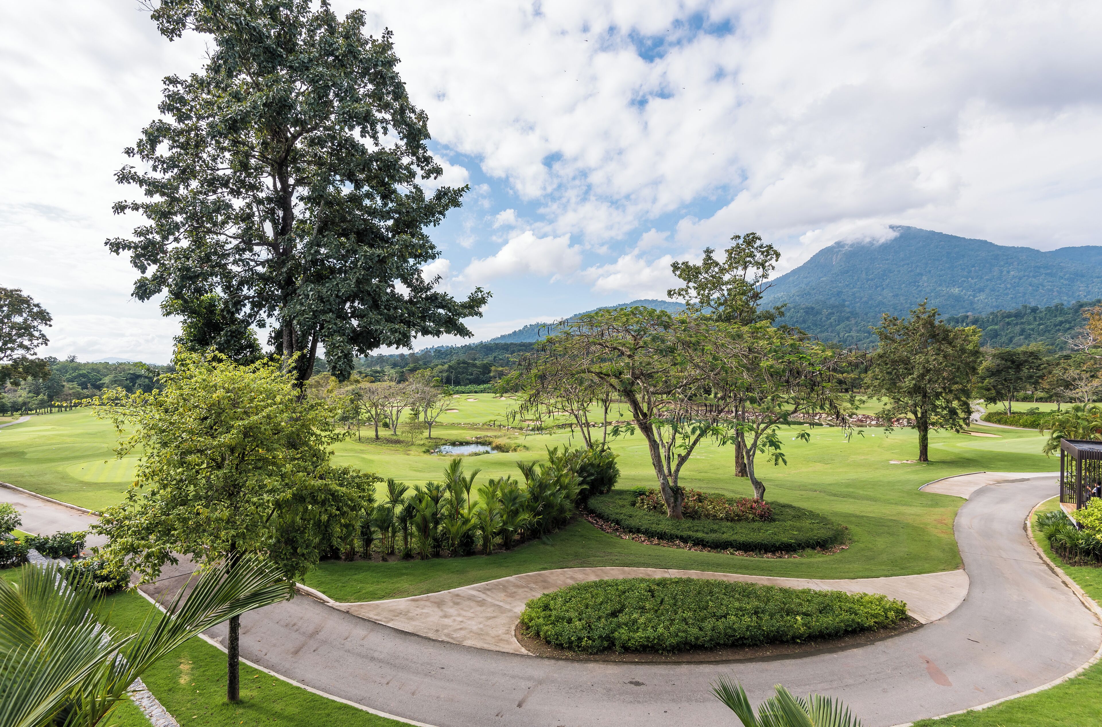 A golf course with a curved road, lush greenery, and a mountain in the background.