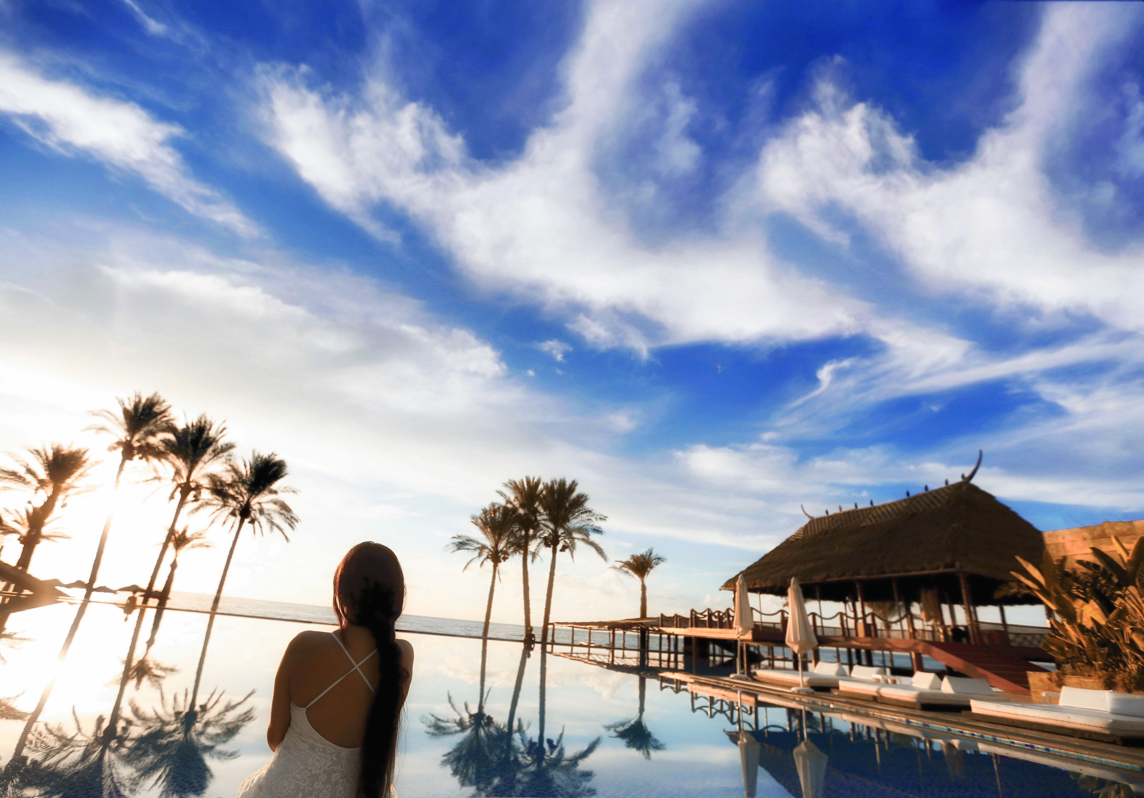 A pool area with palm trees and a thatched-roof structure.