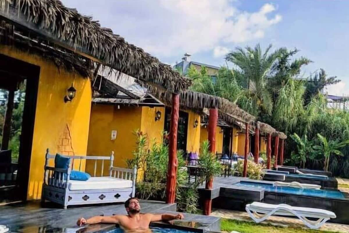 Un hombre descansando en una piscina en un resort con estructuras de techo de paja y palmeras.