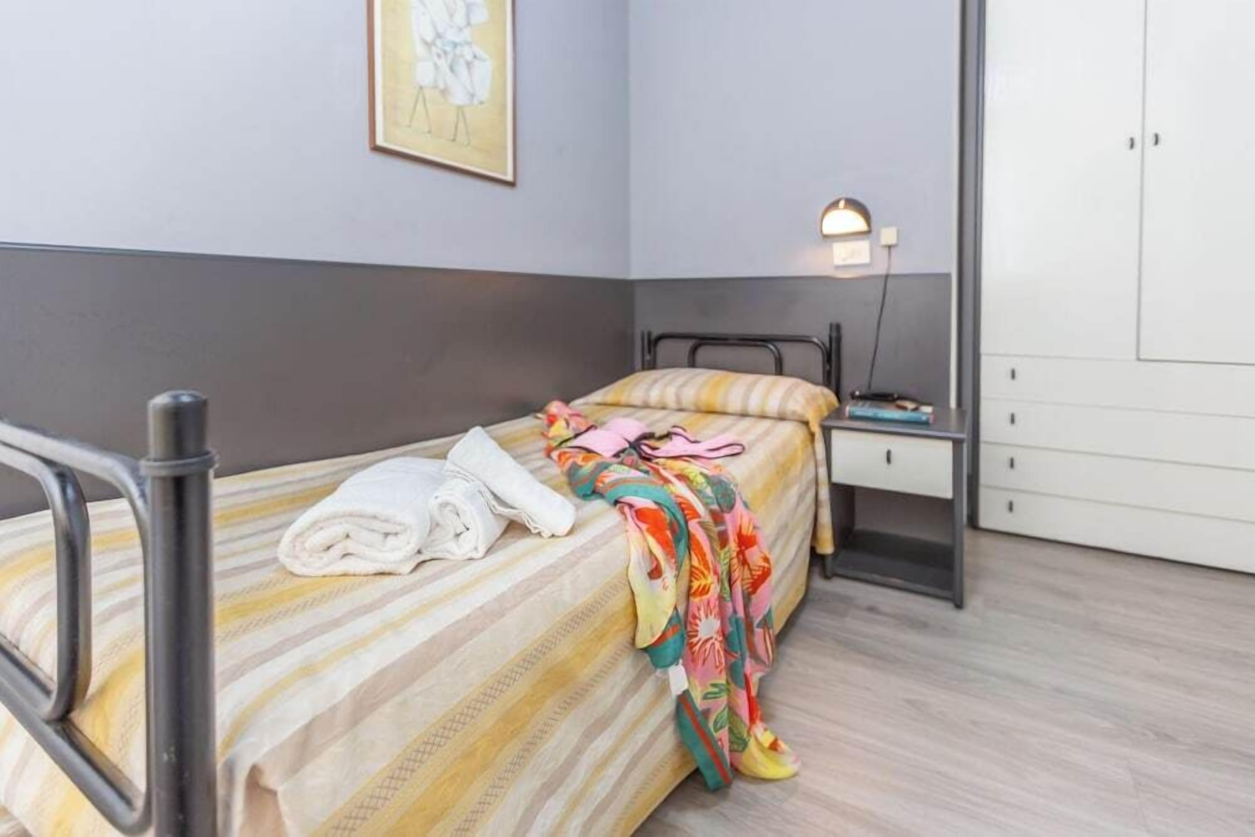 Een eenpersoonsbed met een gestreept beddegoed, een nachtkastje met een lamp, en een kledingkast in een kamer met lichte wanden en houten vloer.