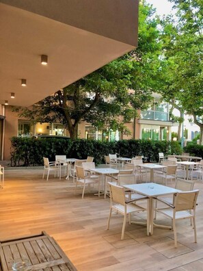 Een terras met tafels en stoelen, omgeven door groen en een gebouw op de achtergrond.