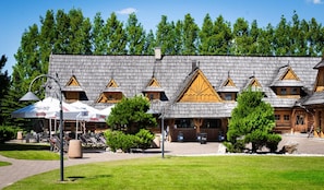A wooden building with multiple windows and a sloped roof, surrounded by trees and outdoor seating. - Hotel Panorama (Mszczonow)