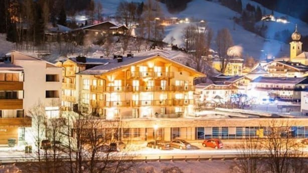 A multi-story hotel with balconies, a parking area, and a view of a snow-covered mountain. - Appart Residence Kristall by Skinetworks (Saalbach-Hinterglemm)