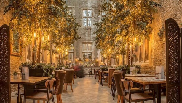 A restaurant with a brick wall, wooden tables, and chairs, decorated with hanging plants and lights.