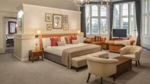 A spacious hotel room with a large bed, a sofa, a chair, and a TV. There are multiple windows providing natural light and a view of the outdoors.