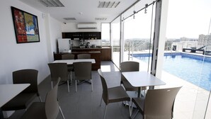 Un espace de repas moderne avec des tables et des chaises, une cuisine en arrière-plan et une vue sur la piscine à travers de grandes fenêtres.