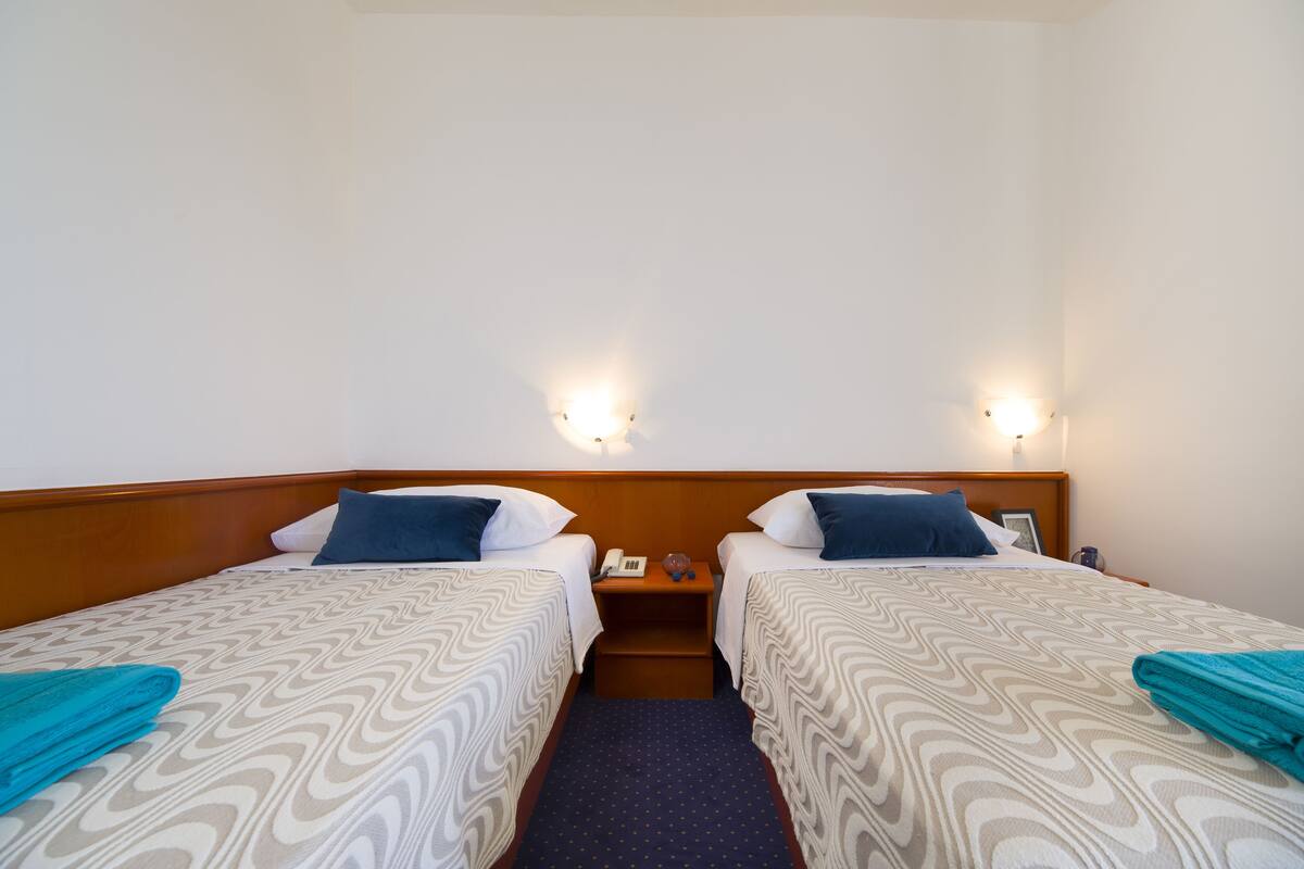A hotel room with two single beds, each with a patterned bedspread and pillows, a wooden headboard, a nightstand with a phone and a glass, and wall-mounted lights.