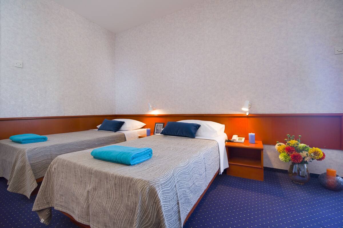 A hotel room with two beds, a nightstand, a vase with flowers, and a blue carpet.
