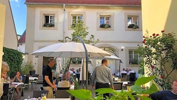Ein Essbereich im Freien mit Tischen und Sonnenschirmen, umgeben von Gebäuden mit roten Ziegeldächern.
