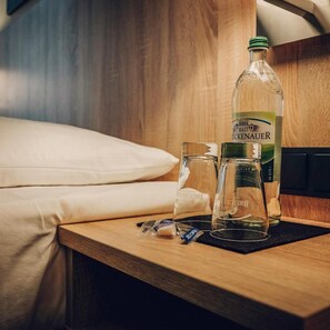 A wooden bedside table with a bottle of Bad Münchner beer, two glasses, and a small packet of snacks.