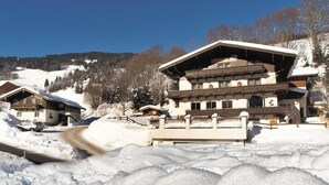 A large, multi-story building with a sloped roof, surrounded by snow-covered terrain and trees. - Pension Gabi (Saalbach-Hinterglemm)
