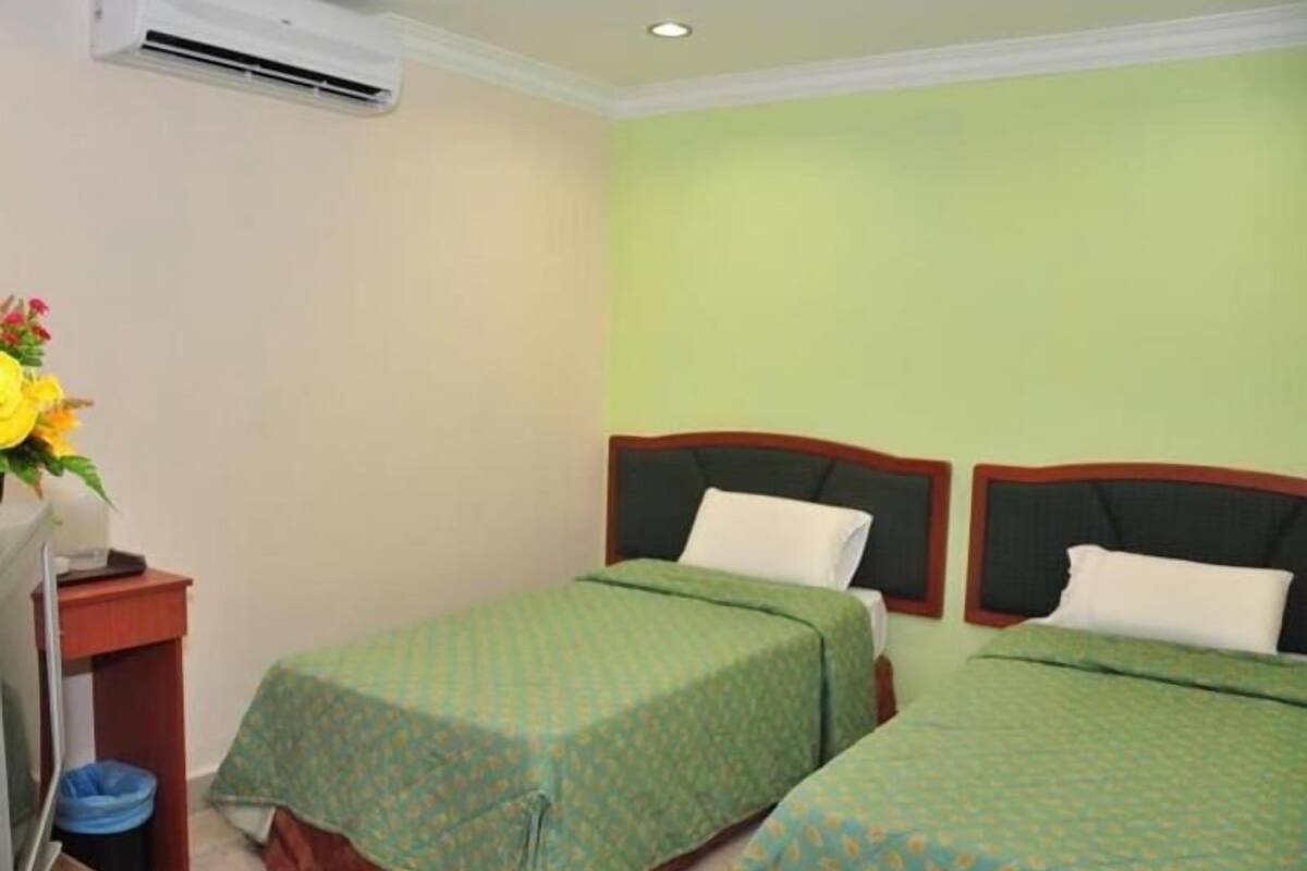 A hotel room with two single beds, a small desk with a chair, a trash bin, and an air conditioning unit on the wall.
