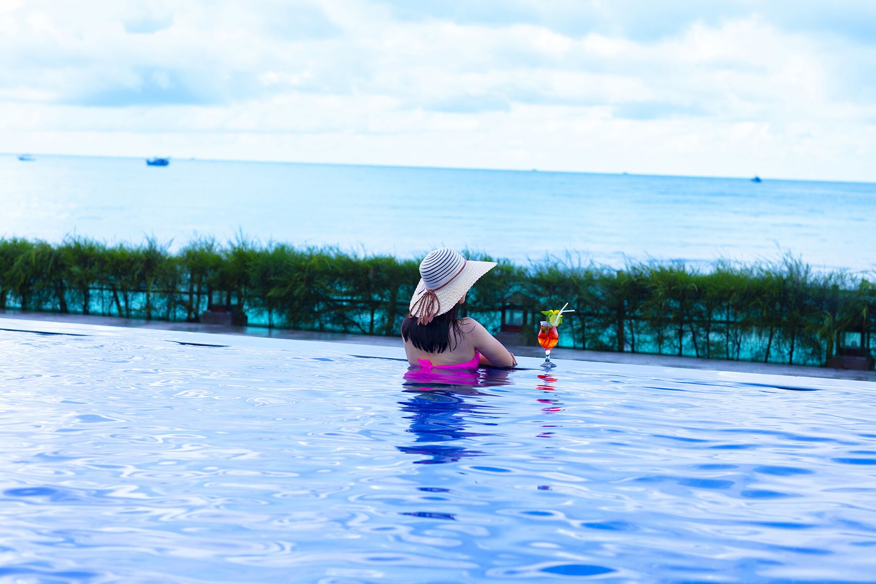A person relaxing in a swimming pool with a drink on the edge, overlooking a body of water and greenery.