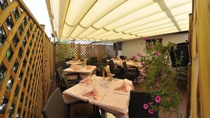 An outdoor dining area with tables set for two, covered by a canopy, and surrounded by wooden lattice and foliage. - Hotel Vela (Trento)