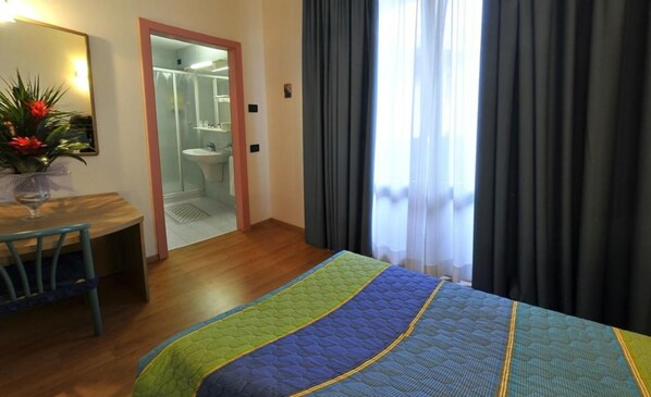 A hotel room with a bed, a desk with a chair, a bathroom visible through an open door, and a window with curtains.