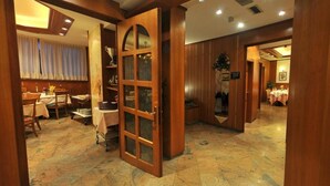 A hotel lobby with a wooden reception desk, a dining area with tables and chairs, and a large wooden door with glass panels. - Hotel Vela (Trento)