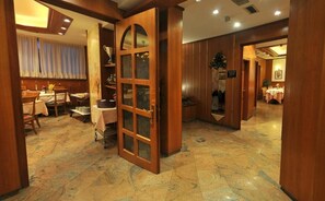 A hotel lobby with a wooden reception desk, a dining area with tables and chairs, and a large wooden door with glass panels.