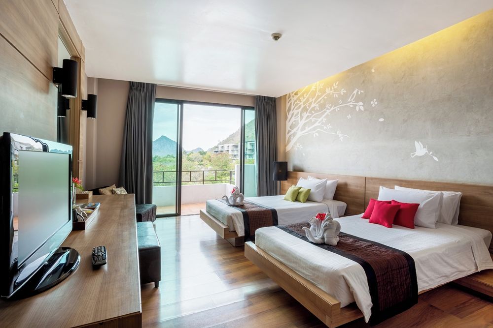 A hotel room with two beds, a TV, and a balcony with a view of greenery and buildings.