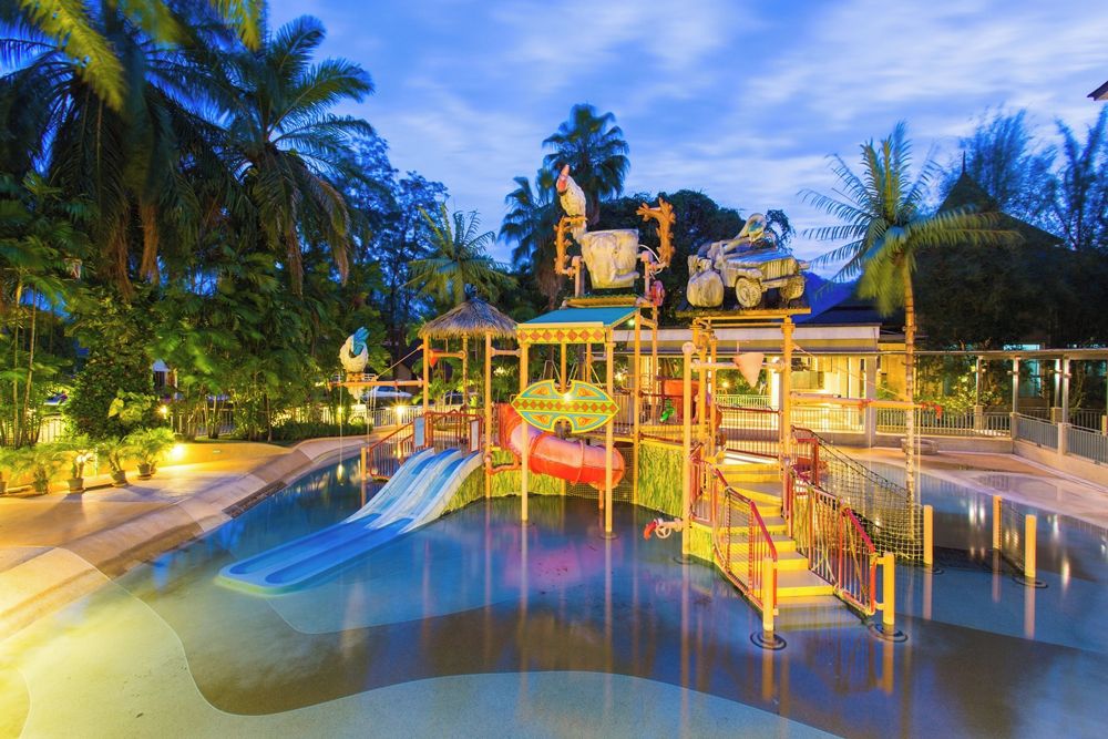 A water park with a central water slide, a play structure, and surrounding palm trees.