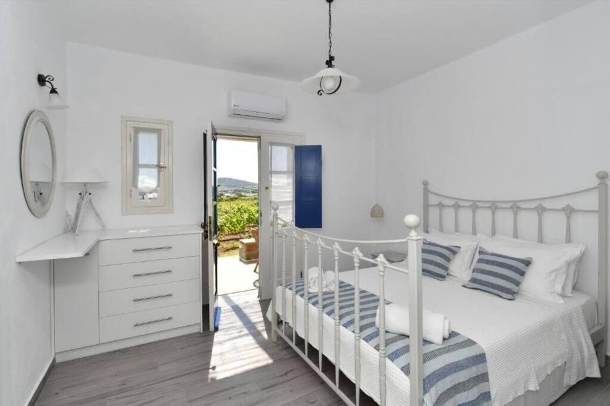 A bedroom with a white bed, a bedside table, a mirror, and a view of the outdoors.