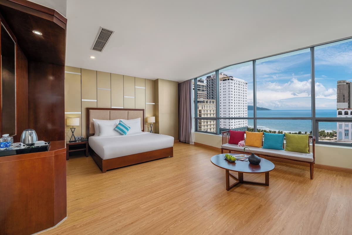 A hotel room with a large bed, a desk, a round table, and a view of the city and sea.