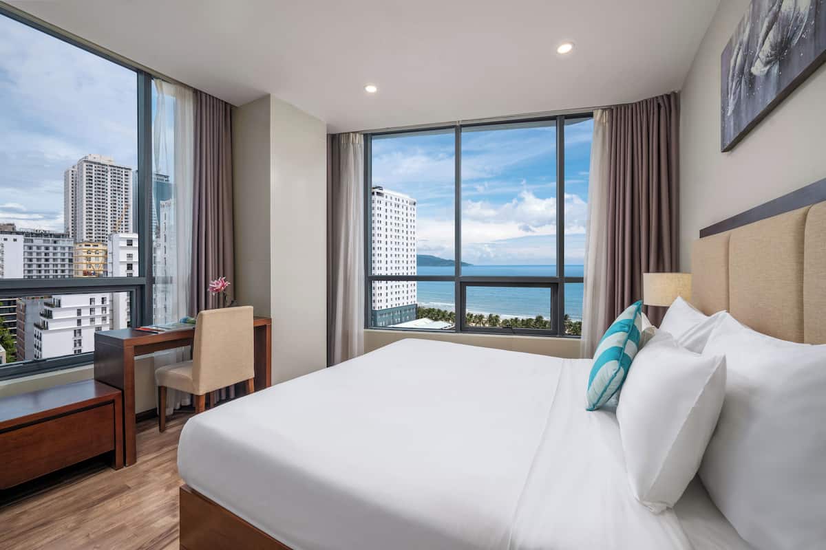 A hotel room with a large bed, a desk, and a chair. There is a window with a view of buildings and the ocean.
