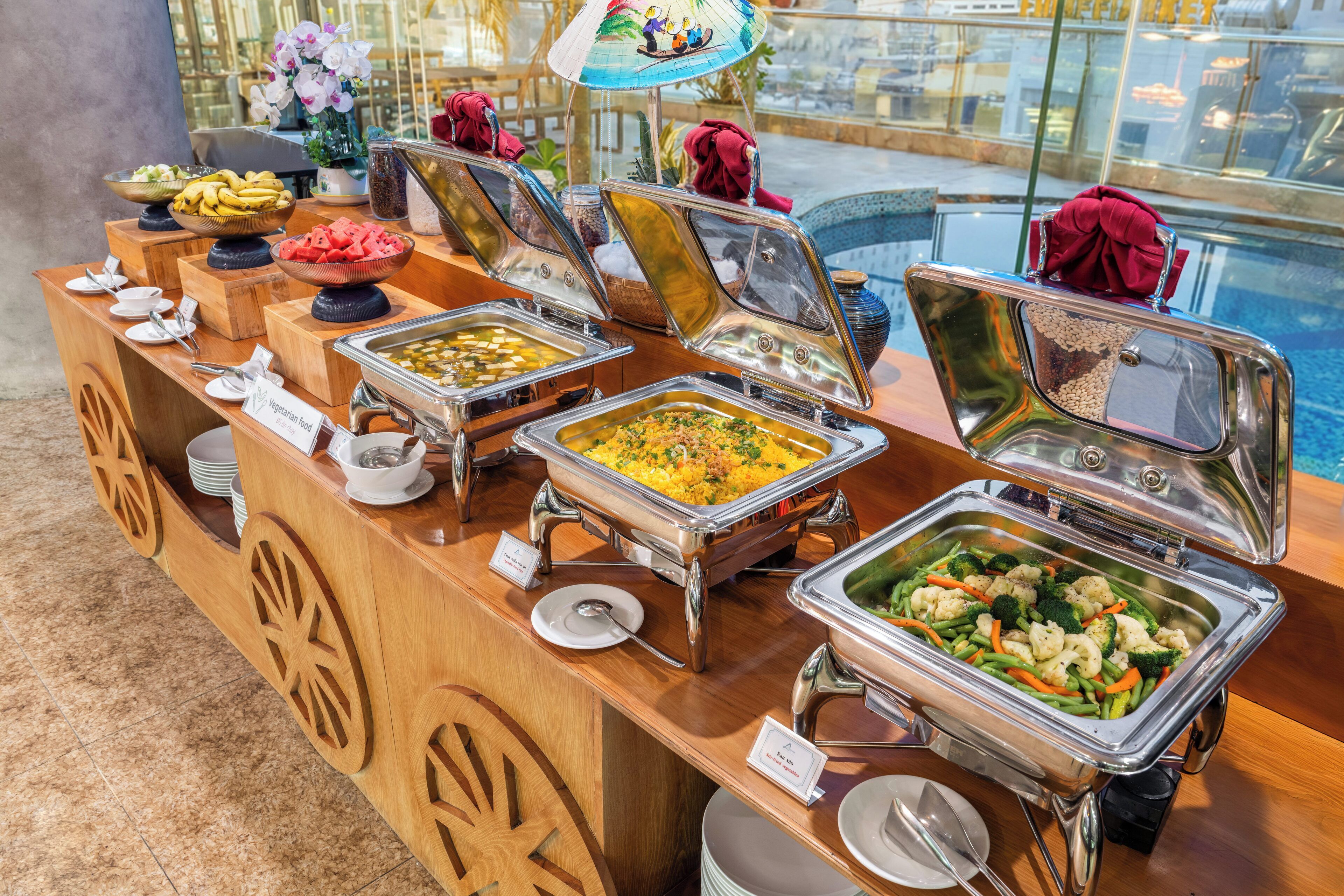 Ein Buffet mit verschiedenen Speisen, darunter Obst, Gemüse und Getreide, präsentiert auf einer Holztischplatte mit Blick auf einen Pool im Hintergrund.