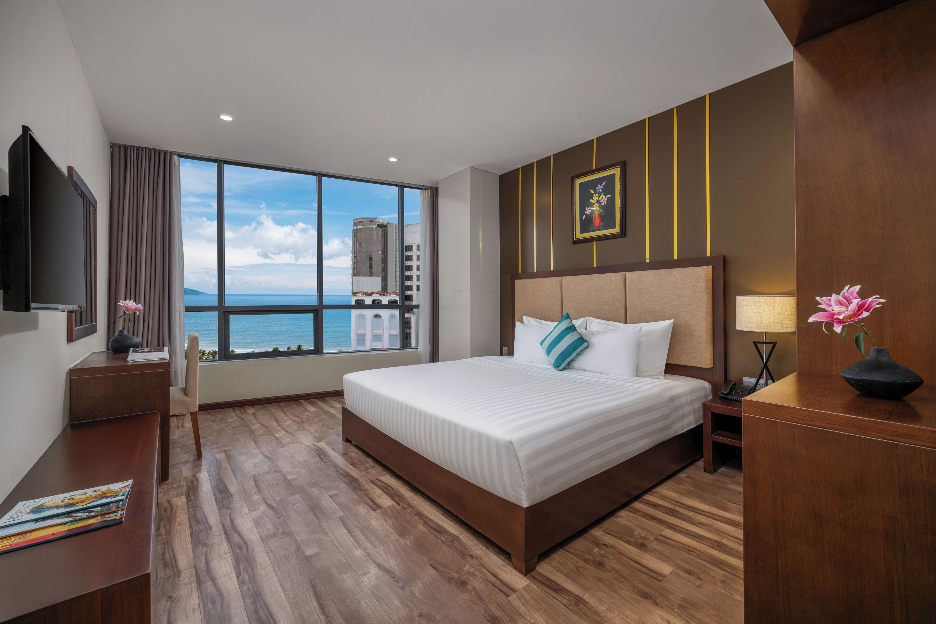 A hotel room with a large bed, a desk, a chair, a TV, and a view of the city and sea.