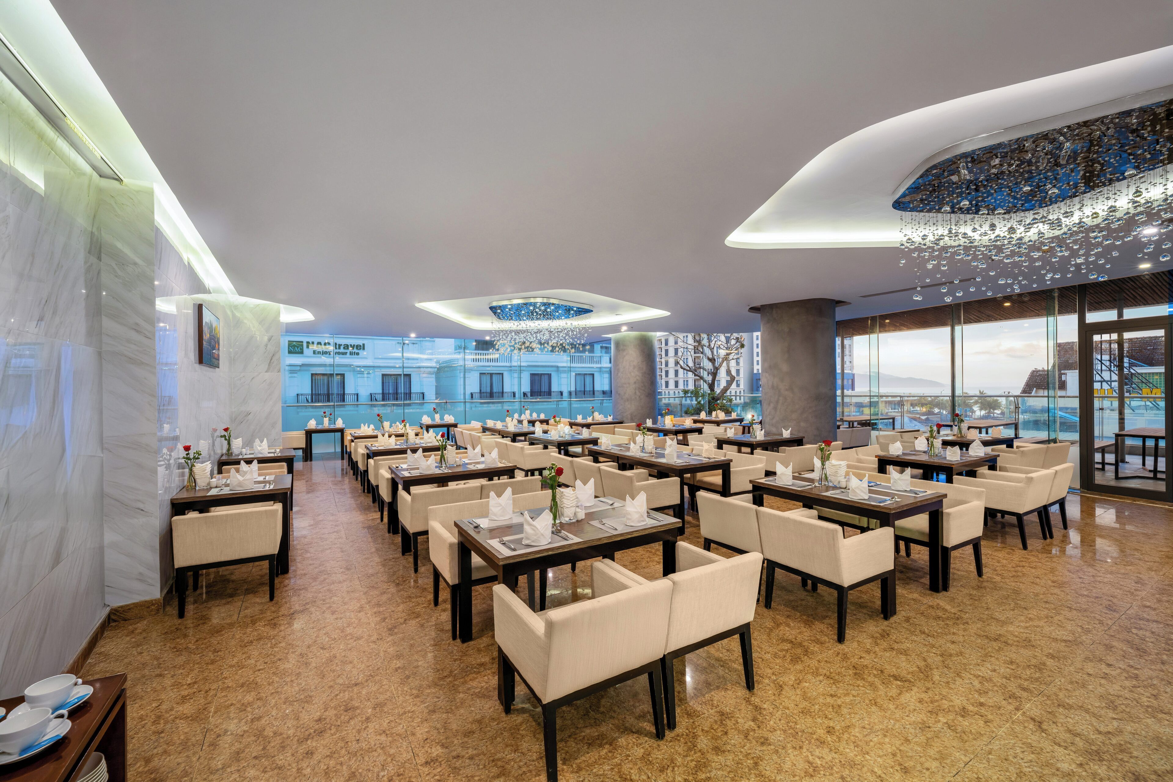 Un vaste espace dédié à la salle à manger, avec de nombreuses tables et chaises, de grandes fenêtres offrant une vue sur les bâtiments et un lustre moderne.