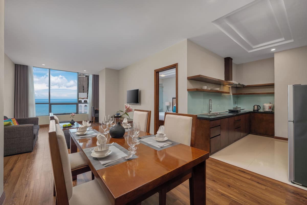 A modern hotel room with a dining area, kitchen, and a large window overlooking the sea.