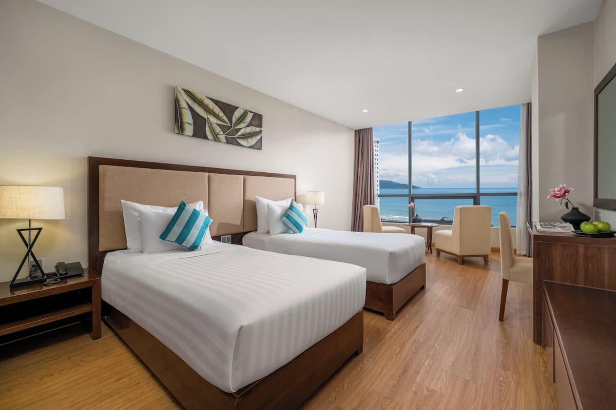 A hotel room with two beds, a desk, a chair, and a large window with a view of the sea.