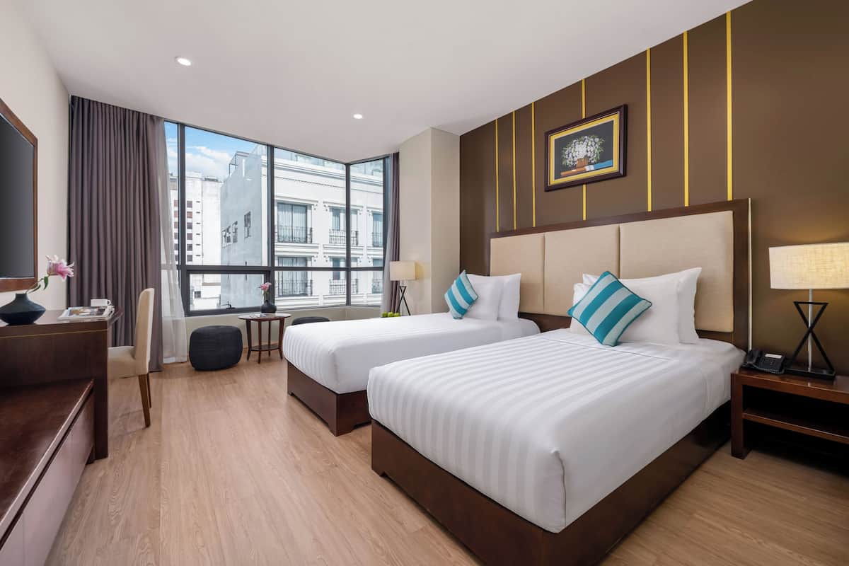 A hotel room with two beds, a desk, a chair, a lamp, and a window with a city view.