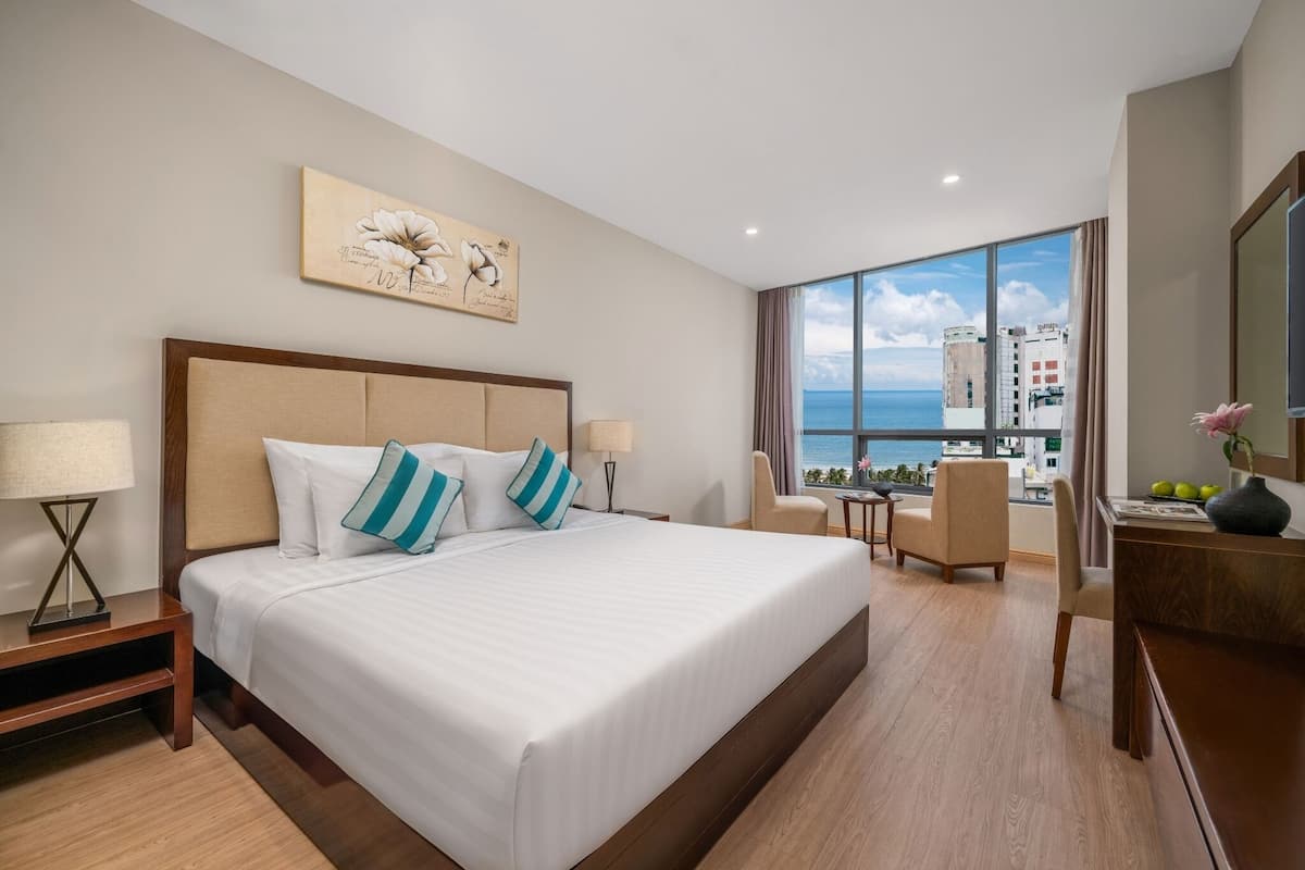 A hotel room with a large bed, a desk, a chair, and a view of the sea.