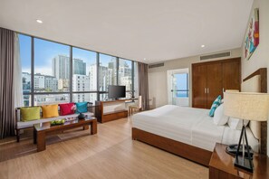 A hotel room with a large bed, a desk with a TV, a sofa with colorful cushions, and a view of the cityscape through large windows. - Avatar Danang (Da Nang)