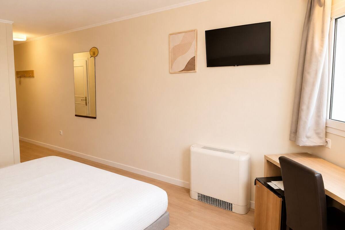 A hotel room with a bed, a desk, a chair, a radiator, a TV, and a mirror.