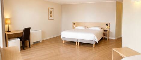 A hotel room with a bed, desk, chair, bedside table, and a lamp.
