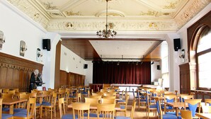 A spacious hall with wooden chairs and tables, ornate ceiling, and a stage with curtains. - Hotel Neustädter Hof (Titisee-Neustadt)