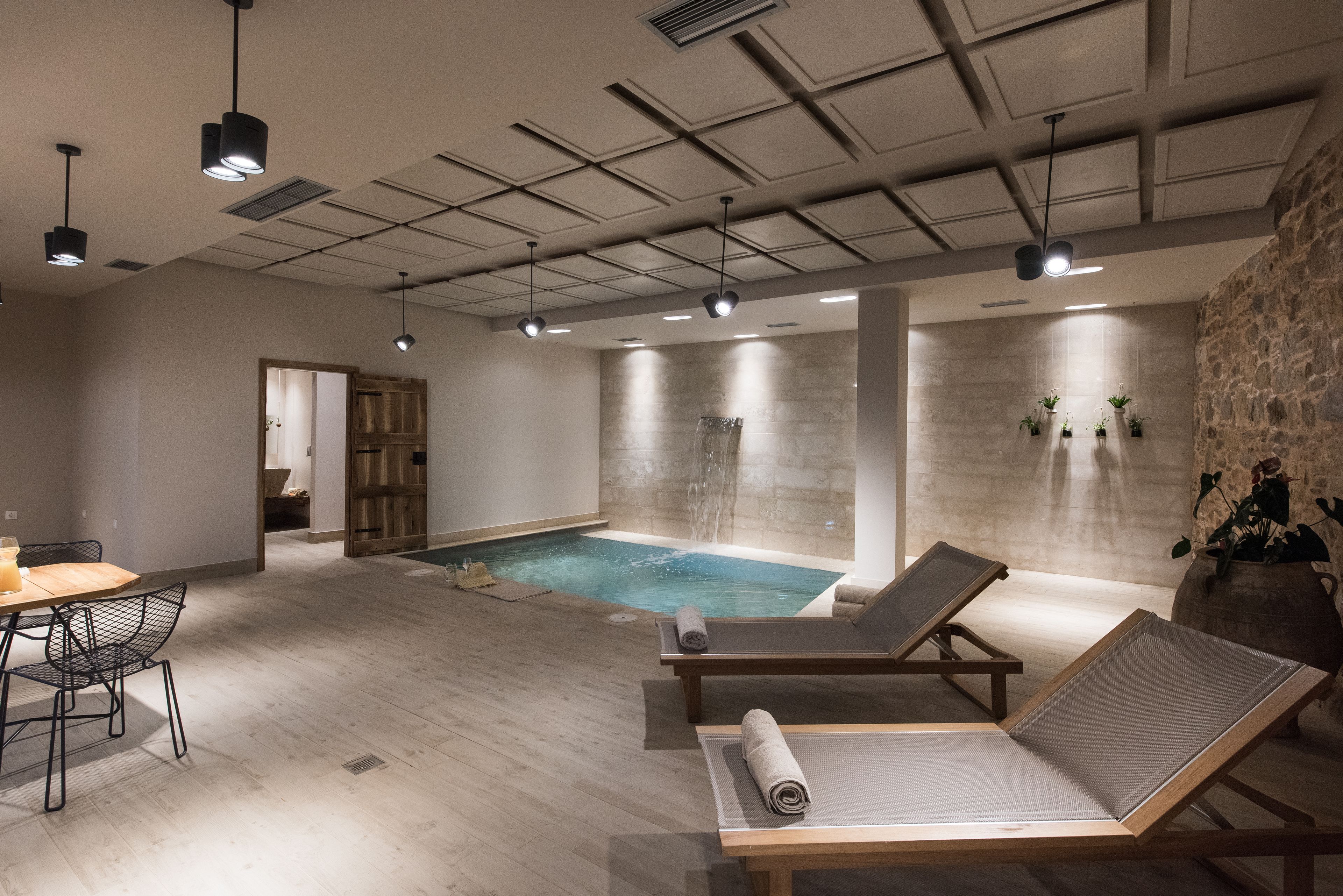 A modern indoor pool area with lounge chairs, a wooden table, and a stone wall.