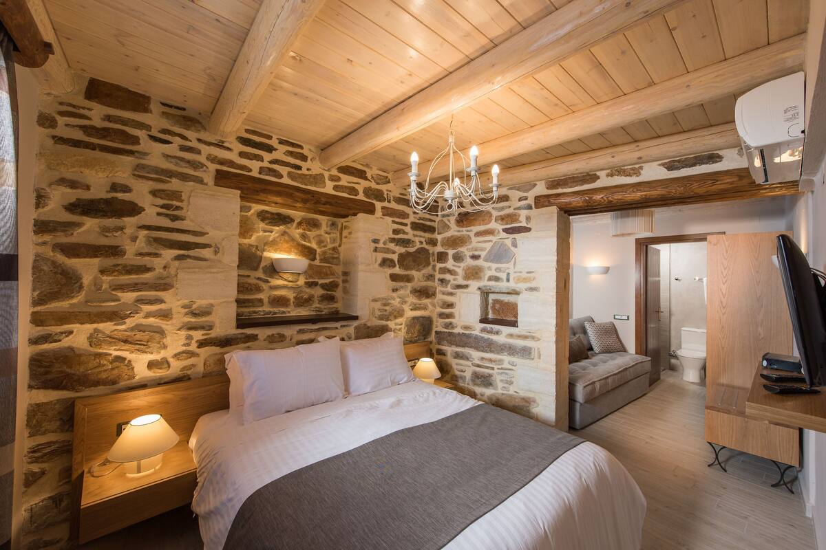 Een gezellige kamer met een stenen muur, houten balken in het plafond, een bed met wit beddengoed, een bedlampje en een kleine zithoek met een bank.