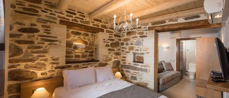 A cozy bedroom with a stone wall, wooden ceiling beams, a bed with white linens, a bedside lamp, and a small seating area with a sofa and cushions.
